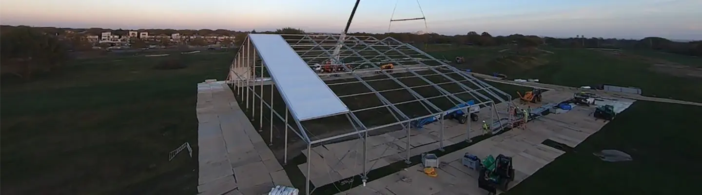 Una grúa instalando un panel de techo en una gran estructura de tela temporal al atardecer en un campo abierto