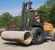 Front angled view of an orange CASE forklift carrying a concrete pipe. A worker is operating on an outdoor construction site.