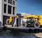 Side view of a yellow JCB telehandler with a forklift attachment moving construction materials on an outdoor worksite.