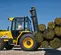 Side view of yellow JCB forklift lifting hay bales with an operator in an enclosed cabin outdoors in a farm setting.