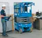 Rear view of a retracted blue Genie scissor lift with an operator standing next to it using the control panel indoors.