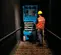 Front view of a blue scissor lift with safety railings and an operator standing in front of lift in a narrow indoor corridor.