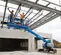 Side view of two workers in safety gear on an extended blue Genie boom lift, elevated under construction roof.