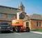 Rear view of an extended orange and beige JLG boom lift with extended arm, in use near a brick building.