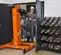 Front angled view of an orange and black vertical mast lift with operator moving it through a doorway.