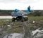 Rear view of a blue and gray telescopic boom lift with tracks, an operator on the platform, working on muddy terrain outdoors.