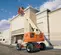 Side view of an orange and beige boom lift with extended arm and worker in basket, in use near building facade.